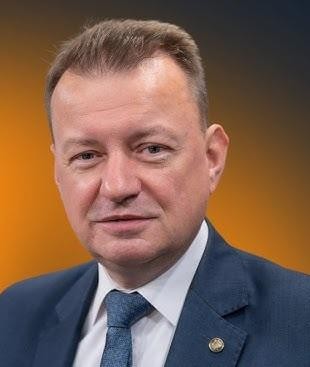 Mariusz Błaszczak jako Minister Obrony Narodowej, stojący na tle nowoczesnego sprzętu wojskowego, symbolizujący jego rolę w obronności kraju.