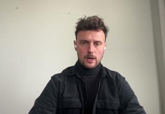 Łukasz Grabarczyk uśmiechnięty, w casualowym stroju, w tle niewyraźne, nowoczesne wnętrze.