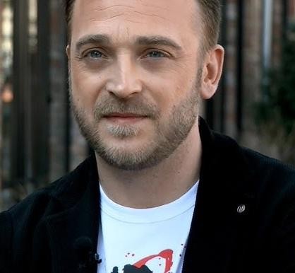 Mateusz Gessler jako juror w programie kulinarnym, w tle elementy studia telewizyjnego.