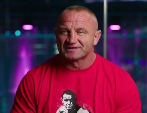 Mariusz Pudzianowski trenujący w siłowni, skupiony na wysiłku, z widocznymi mięśniami i determinacją.