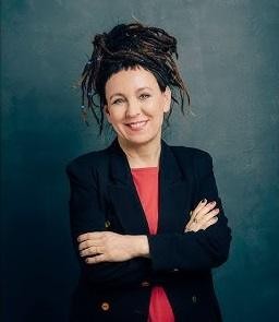 Olga Tokarczuk podczas spotkania autorskiego, podpisująca książki i uśmiechająca się do czytelników, w tle regały z książkami.