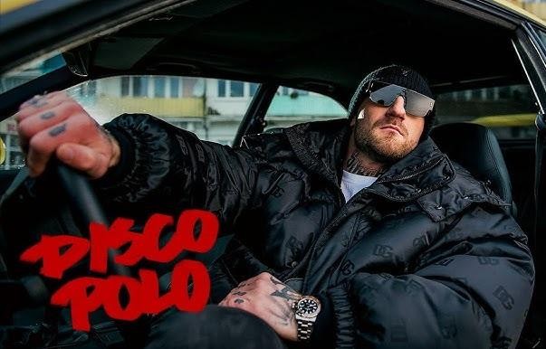 Kizo, polski raper, podczas występu na scenie lub w studiu nagraniowym, otoczony sprzętem muzycznym, z subtelnymi elementami nawiązującymi do jego działalności biznesowej.
